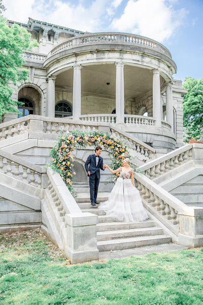 couple on the stairs of the Elkins Estate with the bride looking back at the groom as she extends her hand to him