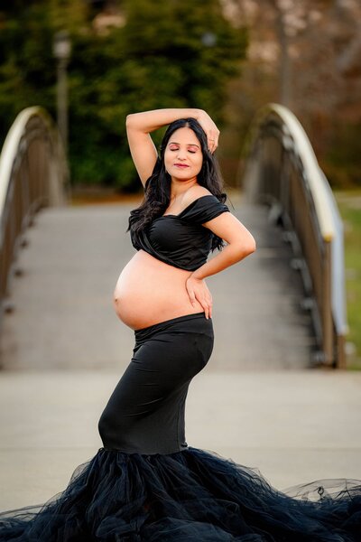 Charlotte Area mother posed in a dramatic way standing in front of a bridge with her belly exposed her arm over her head and her other hand on her back hip dressed and a black maternity dress