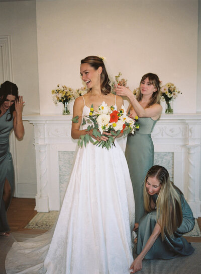Bride in bridal suite holding her bouquet while her bridesmaids adjust her dress