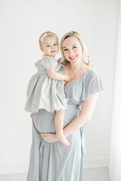 Pregnant mother smiles for portrait in blue flowing dress while holding her toddler daughter on her baby bump