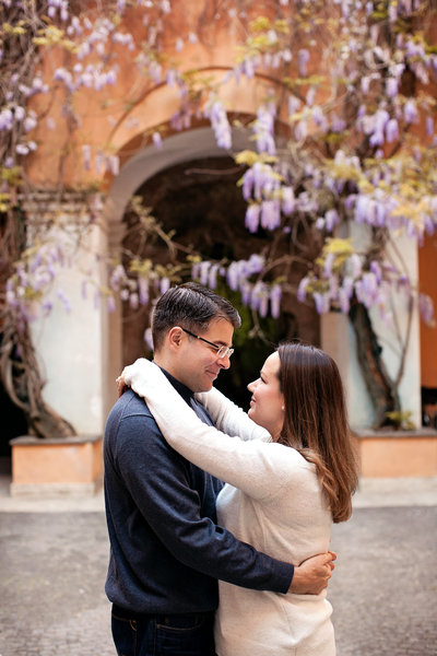 A couple embraced in a piazza with Wisteria in the background. Taken by Rome Vacation Photographer, Tricia Anne Photography