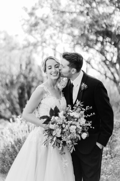 Groom embraces bride and kisses her on the forehead in a botanical gardens