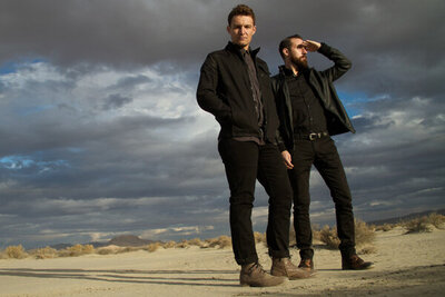 Portrait of music duo Polarcode standing in desert one member shielding eyes with hand big sky with clouds behind them