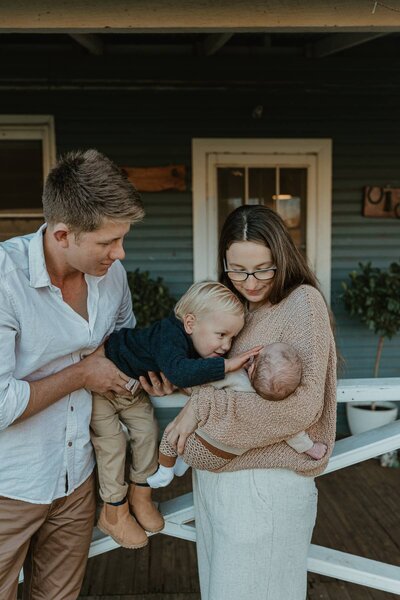 In-home family photography session capturing a toddler kissing their newborn sibling while parents look on.