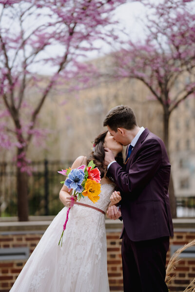 A couple kissing on their wedding day while one of them holds a bright colored bouquet.