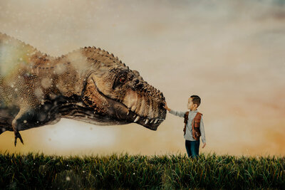 A fantasy photo of a young boy reaching his arm out to touch the nose of a t-rex dinosaur