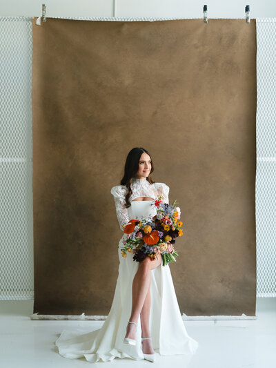 Portrait of bride sitting on a stool with her legs crossed and holding a colorful bouquet with a brown studio background
