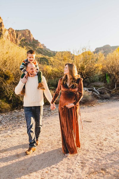 pregnant mom holds her tummy and walks alongside her husband and son on dads shoulders through the Phoenix mountains during glowing sunset