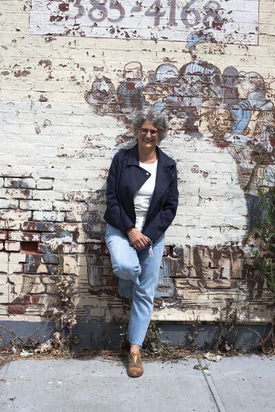 Smiling Amy Posner, a business coach, in casual clothes leaning against a wall with faded murals.