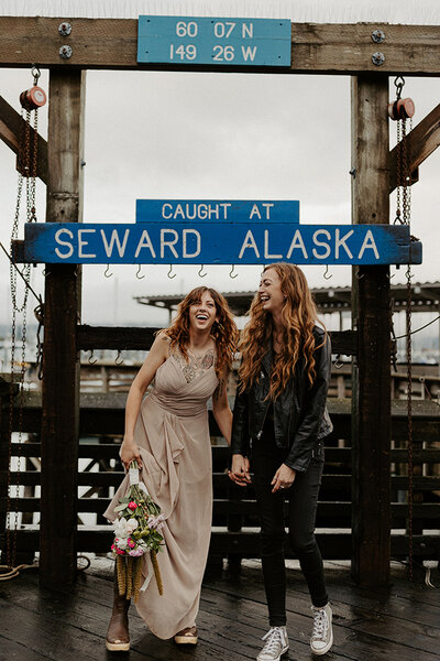 Two women eloping at the Seward, Alaska harbor are standing in front of the infamous "caught in Seward Alaska" sign.  They are holding hands and laughing.