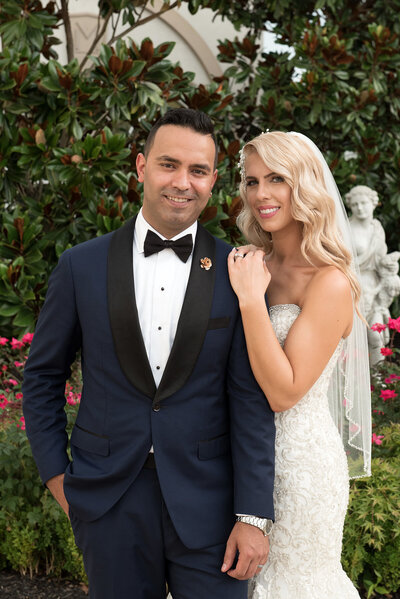 Classic and timeless wedding photo of bride and groom  in a rose garden at a winery with a greek statue in the background. The groom in a handmade fitted blue tux and the bride looking like a 1950 's movie star with bold eyeshadow and lipstick and long wavy blond hair.