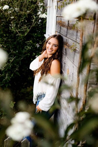 charlotte grad wearing a white top leans against a wall with flowers in the foreground