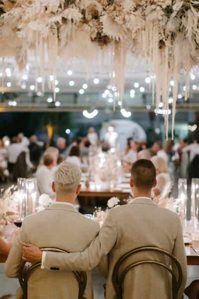 Photo taken from behind two grooms sitting at the head table of their wedding