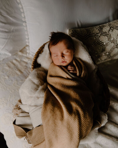 Sweet vintage looking, dark aesthetic newborn photoshoot by Morgan Ashley Lynn Photography in Milwaukee, WI of a baby boy wrapped up in a brown blanket with one hand sticking out near his face, sleeping. He is laying on a soft bulky white blanket and on a bed with boho looking pillows