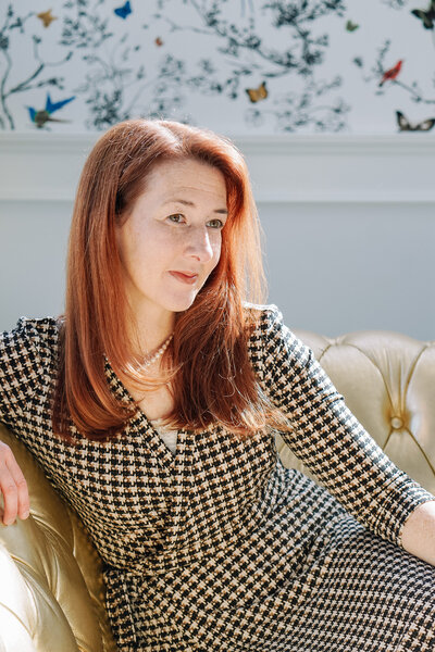 Portrait of owner and photographer, Carolyn Ann Ryan, wearing a black and cream checkered dress sitting on a couch with sunlight behind her.