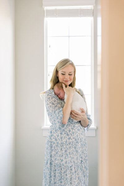 Image of new mother holding baby standing near window in blue dress taken by Sacramento Newborn Photographer Kelsey Krall