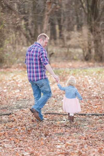 Father in plaid shirt walks through a field of leaves holding his young daughters hand
