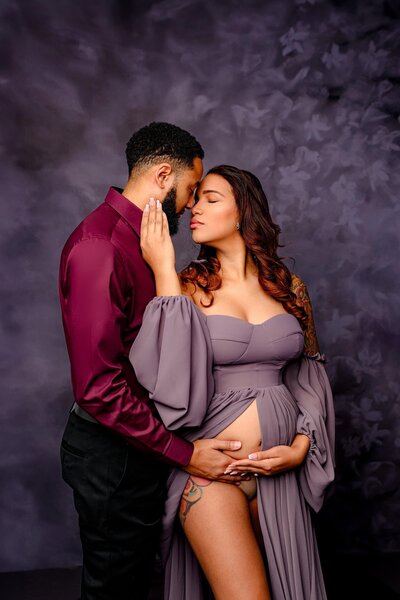 mom gently leans back against dad touching his face as they are nose to nose in their studio maternity session.  mom is wearing a dusty purple and dad is wearing red shirt and pants