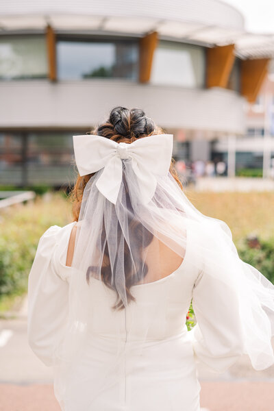 A bride stands with her back to us facing the gemeentehuis and her curled hair, veil, oversized bow, and scoop neck wedding dress details are visible