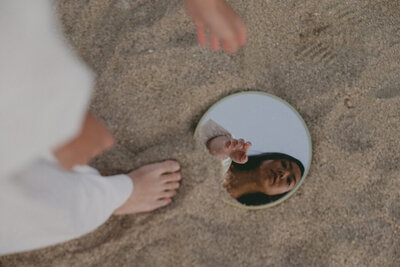 A woman looking down at a mirror in the sand. Representing someone who would benefit from therapy for self-esteem and confidence counseling in Brooklyn, NY.  Start setting and reaching self-esteem goals with support of a confidence counselor in NYC.