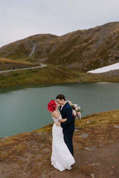 A couple in wedding attire hug and embrace each other in front of Summit Lake atop Hatcher Pass in Alaska during their adventure elopement.