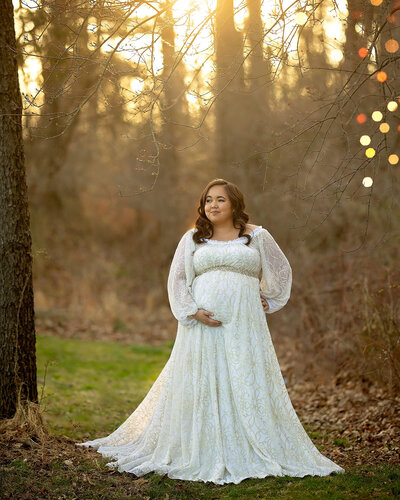 A beautiful dark haired plus sized pregnant woman wears a white dress in the woods, holding her belly