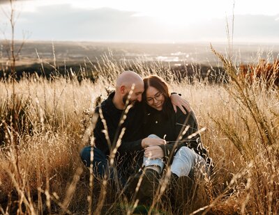A couple is sitting on a mountain surrounded by tall brown grass. A city can be seen behind them.