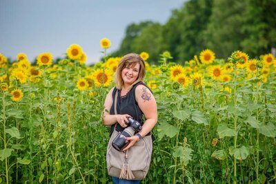 A woman holding a camera and camera bag laughing with a sunflower field behind her