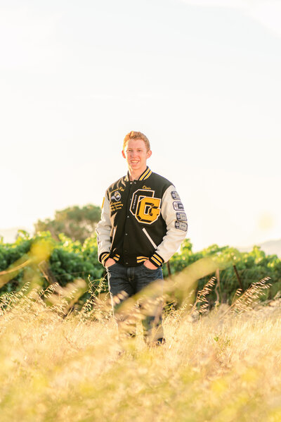High school senior guy wearing a varsity jacket in a field of yellpw grass
