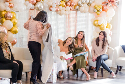 A woman opens a gift in a room adorned with a balloon arch, captured perfectly by an event photographer in Fayetteville, NC. She is seated among three other women, with one standing nearby on light-colored chairs. The atmosphere appears festive and relaxed.