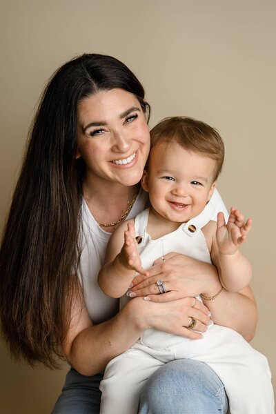 mother dressed in jeans and a white shirt cuddles her one year old son on her lap.  he is dressed in white overalls
