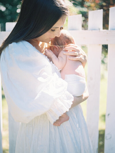 Mom in white dress holding newborn in front of a white fence on film by richmond newborn photographer