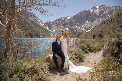 A groom in a suit sits on a rock next to a lake while his bride stands next to him in a white dress.