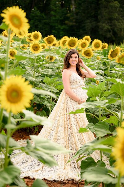 Expectant mother tucks her hair behind her ear and cradles her baby bump while standing in a charlotte area sunflower field wearing a boho dress