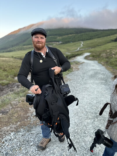 Man in black shirt and hat holding camera gear on a gravel path in a green, hilly landscape—perfect inspiration for a Switzerland photo retreat in the Alps for adventurous photographers.