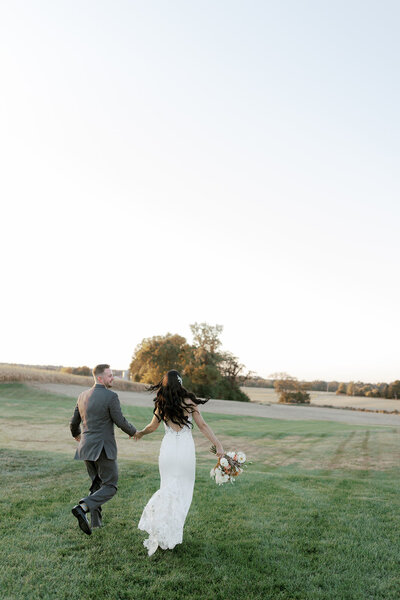 A couple holds hands and runs away towards a field at sunset on their wedding day. The bride is holding a bouquet in one hand and her dress is bustled as the groom looks towards her smiling.