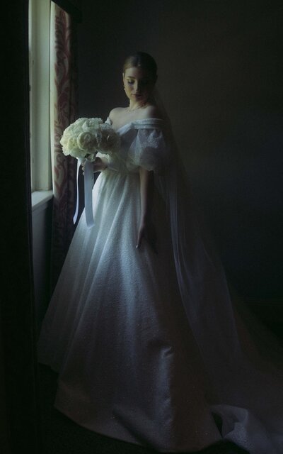 Bride holding her bouquet by the window, bathed in soft natural light, capturing a serene and intimate moment before her wedding day begins