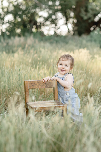 baby boy in blue overalls holding on to chair in tall grass