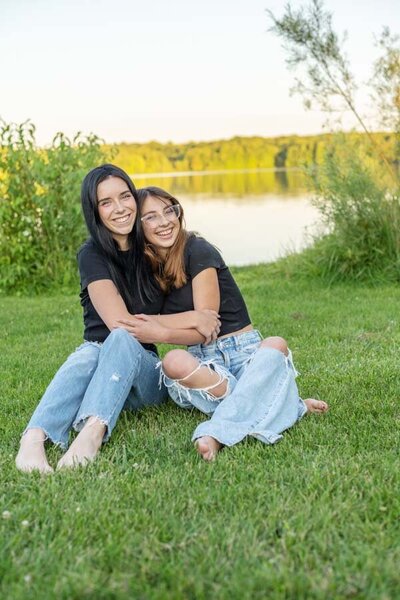 two woman sitting on the grass in front of a lake hugging