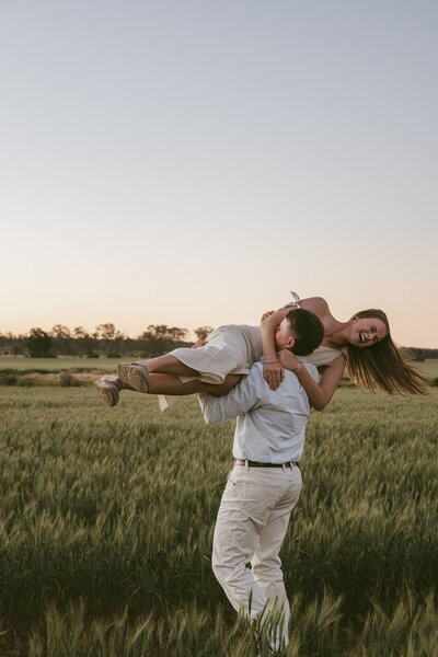 A playful moment during an engagement photoshoot, with a man lifting his laughing partner in an open field at golden hour.
