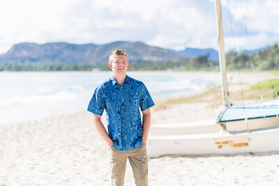 senior boy standing on a sandy beach with his hands in his pockets with a boat in the background.