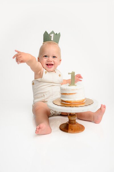 little boy sits wearing a green crown playing in his first birthday cake