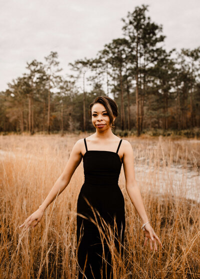 young black woman wearing black dress trails fingers on tall brown wheat grass around her while smiling at photographer brittney stanley of panama city fl