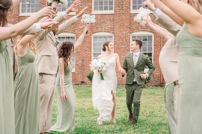 A happy couple running through the hands of their wedding party in front of a warm brick wall in July by JoLynn Photography