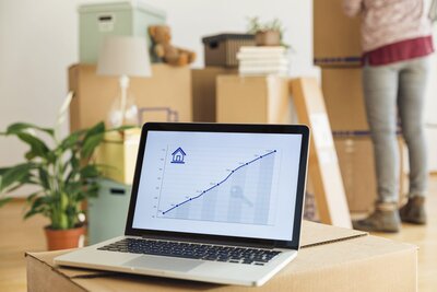 a laptop showing the growth of a real estate investment with a woman fixing boxes for her new property in the background