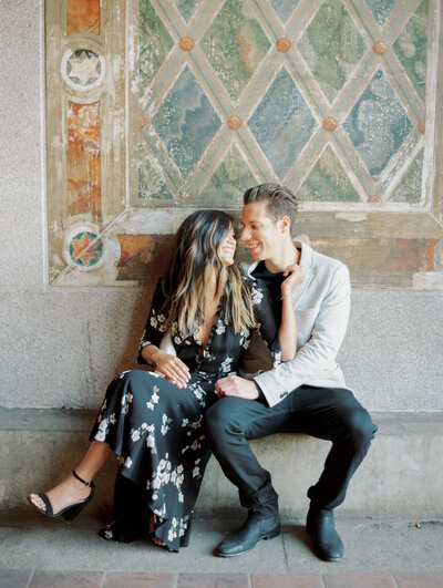 Engagement photography of a man and women sitting side by side on a cement step, in front of an old Italian style blue and orange argyle pattern painted cement wall.