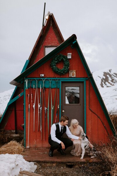 An eloping couple crouch next to their dog in front of the infamous red cabins atop Hatcher Pass in Alaska during their snowy May elopement.
