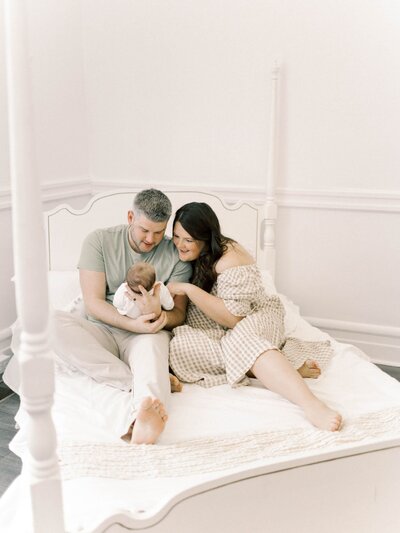 Man and woman site on white frame bed in a white lit room, holding their newborn baby.