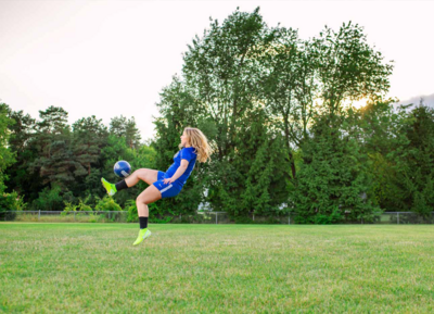 high school senior girl kick soccer ball in the air with green trees in the background