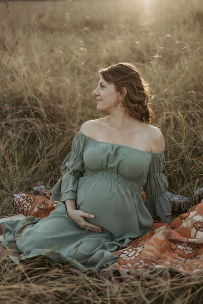 Pregnant mama in strapless green dress cradling her belly while sitting in a grassy field with the sun glowing on her